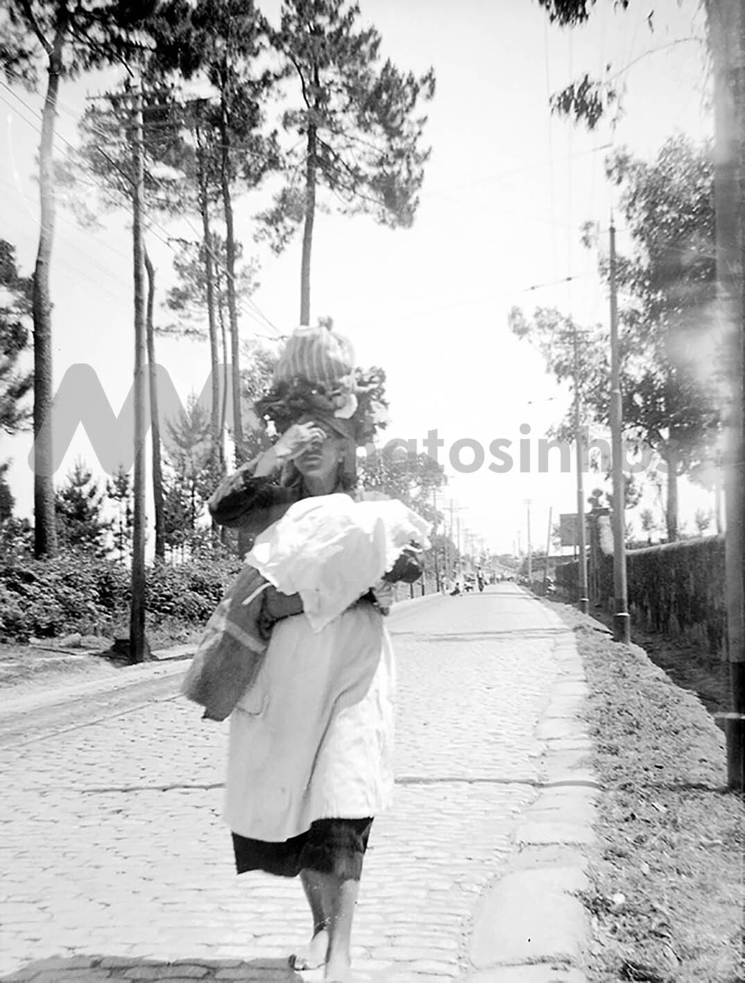 Mulher na rua com a trouxa &agrave; cabe&ccedil;a e crian&ccedil;a ao colo, em S. Mamede de Infesta